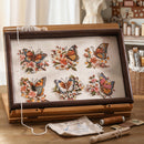 A wooden embroidery frame holding six colorful butterfly and floral designs on cream fabric, shown on a tabletop with thread, a small pouch, and other crafting supplies in a softly styled room.