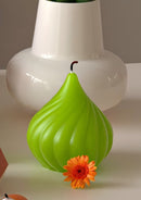 A bright lime green pear-shaped candle with a ribbed texture and a small dark stem, styled in front of a white ceramic vase with an orange flower at its base.