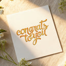 White card with 'congrats to you' in gold text on a wooden surface with flowers.