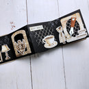 A vintage-style pop-up book or album spread displayed diagonally on a white wooden surface. The black textured pages feature illustrated inserts including a lamp, an angel, a coffee cup, a dress, and Polish text labels.