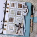A styled flat-lay of a light blue leather-bound planner or journal, open to a decorated page with vintage postage-stamp frames, script writing, a camera illustration, and a large number 5 tab. A pen with a vintage-patterned barrel rests on the right side against a whitewashed wood background.