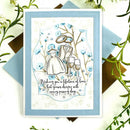 A square layered card with a light blue border and a central illustration of a wedding couple, rings, two champagne glasses, and the phrase 'Wishing you a lifetime of love that grows deeper with every passing day.' The card is styled on a white surface with blue and brown paper backing and green floral sprigs around it.