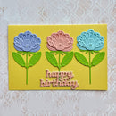 A handmade square greeting card with three embossed paper flowers in blue, pink, and light blue on a yellow background, with 'happy birthday' written in pink script at the bottom.