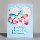 A handmade square birthday card with a light blue background, featuring a layered heart design with a blue and yellow bird on a pink branch and the words 'happy birthday' in light blue script at the bottom.