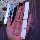 A hand holds three narrow printed strip templates or labels in pink, white, and silver-gray, photographed over a dark wood surface with a lace cloth and small decorative props in the background.