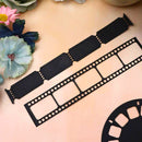 Black metal film strip cutouts, including a full-length strip and a partial reel section, arranged on a peach-colored surface with decorative flowers in the corner.