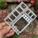 A hand holds a silver metal die-cut frame template with multiple rectangular and square cutouts, set against a rustic background of bark, moss, and dried leaves.
