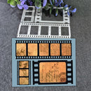 A flat sheet of cardstock or craft paper featuring a vintage film strip design with antique lettering and sepia-toned document illustrations, displayed on a gray textured fabric background with decorative purple flowers and metal film-frame cutters above it.