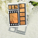 A white rectangular card or plaque featuring vintage film-strip and envelope designs with tan parchment-style inserts and handwritten script, displayed on a white lace surface with white flowers in the background. Silver metal film-frame molds are placed underneath.