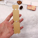 A hand holding a tan kraft paper ticket-shaped craft die-cut template, with other crafting dies and small decorative items on a white textured background.