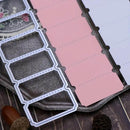 A close-up of a planner or notebook insert set with a white metal die-cut template over pink and white paper tabs on a dark textured surface.