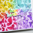 A close-up of a colorful handmade card featuring a rainbow gradient background with illustrated butterflies in red, pink, purple, blue, and yellow. A die-cut sticker reading “happy birthday” is layered over the design.