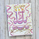 A handmade birthday card featuring a two-tiered cake with purple and pink striped layers, three lit purple candles, pink confetti streamers, and gold foil lettering that reads 'Happy Birthday'. The card is displayed on a light gray, weathered wood background.