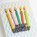 A close-up of a handmade birthday card featuring five colorful striped birthday candles with yellow flames and a black-and-white 'HAPPY BIRTHDAY' text overlay, set against a white card with a yellow and blue polka dot pattern.