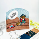 A handmade pirate-themed card featuring a cartoon bear dressed as a pirate, a treasure chest, a red crab, and a speech bubble that reads 'VE LOST A TREASURE'. The card is styled on a white surface with craft dies and a patterned card partially visible in the foreground.