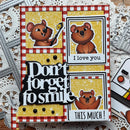 A handmade layered greeting card featuring cartoon brown bears on a yellow glitter background, with red-and-white gingham borders, black-and-white text reading 'Don't forget to smile' and 'THIS MUCH!', plus a photo frame panel that says 'I love you'. The card is photographed on top of white die-cut paper and metal embossing tools.