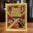 A handmade Harry Potter themed greeting card standing on a wooden table. The card features a yellow border, a large red and brown X background, the phrase 'Don't forget to smile' in gold lettering, and a cute cartoon bear wearing a red and yellow striped scarf and black robe holding a wand.