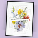 A framed handmade greeting card featuring a watercolor floral design with yellow, blue, and red flowers on a white background with a black frame, set against a light purple wall.