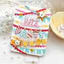 A handmade layered paper card with a 'let's PARTY' design, styled on a white fabric surface with decorative props like a small white flower and a yellow dish.