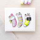 A handmade greeting card featuring three colorful Christmas stockings with candy, gifts, and a teddy bear, with a black label reading 'JOY & CHEER'. The card is displayed on a light beige surface with markers partially visible at the top.