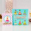 A birthday-themed gift bag and matching card set on a white surface. The bag is white with pink polka dots and a colorful party hat illustration, with the word 'celebrate' in gold script.