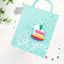 A handmade light blue paper gift bag with a scalloped handle, featuring an embossed floral pattern, a small layered birthday cake die-cut element, and the words 'for you' in white script. The bag is photographed on a white surface with decorative green leaves and small confetti.