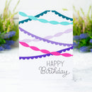 A white square birthday card with colorful decorative ribbon graphics and the words 'HAPPY Birthday' printed on it, photographed standing on a light surface with blurred flowers in the background.