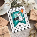 A handmade layered birthday card featuring a white and black paw-print pattern border, a teal and turquoise scalloped center, and a white Dalmatian dog illustration holding an orange balloon. The card is styled on a gray surface with wrapped gift boxes, white flowers, and a cup of coffee.
