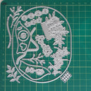 A metal die-cut craft sheet featuring a whimsical tree of life design with branches, leaves, acorns, and a heart-shaped trunk, displayed on a green cutting mat with grid lines.