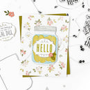 A handmade greeting card featuring a watercolor illustration of a mason jar filled with yellow pickles and the word 'HELLO' in yellow, layered over a white floral background. The card is displayed on top of a green envelope, surrounded by craft embellishments and clear stamps on a white background.