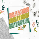 A handmade greeting card with diagonal leopard-print stripes in coral, olive, and mint green, featuring the words 'HEY HI HELLO' and small flowers. The card is styled in a flat-lay arrangement with metal letter cutouts and other crafting tools on a white background.
