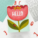 A handmade greeting card featuring a stylized flower with the word 'HELLO' in white letters, surrounded by scattered alphabet stickers on a white background.
