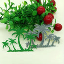 Green and silver tropical palm tree cutout designs displayed on a white background with decorative artificial greenery and red berries behind them.