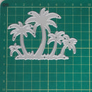 A silver metal die-cut craft stencil featuring a tropical palm tree design, displayed on a green cutting mat with grid lines and centimeter measurements.