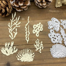 A set of coral-shaped craft cutouts or stamps displayed on a wooden surface, with pine cones and a small decorative object in the background. The left side shows flat cream-colored pieces, and the right side shows white die-cut outlines of matching coral shapes.