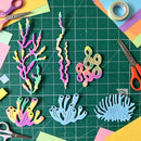 A flat-lay of colorful coral-shaped craft cutouts arranged on a green cutting mat, surrounded by scissors, paper scraps, and tape. The coral pieces are in pastel rainbow colors including pink, yellow, green, blue, and purple, with one larger light blue piece at the bottom right.