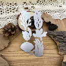 A set of silver metal die-cut craft shapes with botanical and leaf silhouettes, arranged on a rustic wooden surface with a lace doily, pinecones, dried leaves, bark, and a small wood slice.