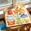 A wooden children's puzzle board with colorful cartoon sea creatures, displayed on a woven stool near a window with a glass of lemonade.