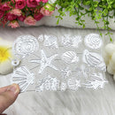 A hand holds a clear acrylic sheet featuring white laser-cut sea urchin, starfish, and shell designs, shown against a soft white fabric background with decorative flowers and greenery.