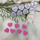 A set of heart-shaped decorative stickers or tags arranged on a light surface with greenery and white flowers. The larger silver-gray hearts read phrases like LOVE, YOU ROCK, XOXO, and ME & YOU, while the smaller pink hearts read LOVE, TRUE LOVE, XOXO, HOT STUFF, and ME & YOU.