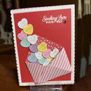 A handmade greeting card with a red background and scalloped white border, featuring the phrase 'Sending Love Your Way' in white script. The card shows a striped red-and-white envelope with pastel heart-shaped pieces and small round heart pieces that read 'YOU ROCK', 'TRUE LOVE', 'LOVE', 'XOXO', and 'ME YOU'.