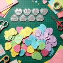 A flat-lay of colorful heart-shaped and cloud-shaped craft stickers on a green cutting mat, surrounded by scissors, paper scraps, and tape. The stickers feature various romantic phrases like 'LOVE', 'TRUE LOVE', 'I LOVE YOU', and 'XOXO' in white lettering.