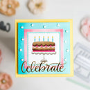 A handmade square greeting card featuring a layered birthday cake illustration, a light blue and pink inner border, a yellow outer border, and the word 'Celebrate' in metallic gold script. The card is displayed on a light gray surface with soft-focus decorative props in the background.