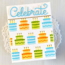 A square white greeting card featuring the word 'Celebrate' in light blue script at the top, with rows of colorful illustrated birthday cakes below. The card is photographed against a soft white background with a faint lace-like decorative element behind it.