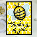 A handmade greeting card featuring a cute yellow and black striped bumblebee with light blue wings, set against a yellow honeycomb pattern. The card has the text 'thinking of you' in black cursive and is displayed on a white surface with blurred craft supplies in the background.