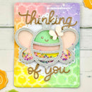 A handmade greeting card or art plaque featuring a cute green avocado character with butterfly ears and the words 'thinking of you' in gold foil, set against a pastel rainbow background on a white wood surface.