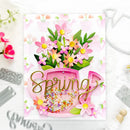 A handmade spring-themed card featuring layered pink and green paper flowers, a pink watering can filled with multicolored beads, and a gold metal 'Spring' script overlay, styled on a white surface with metal dies.