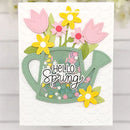 A handmade greeting card featuring a green watering can design with layered pink tulips, yellow flowers, green leaves, and the words 'Hello Spring' in white script. The card is displayed against a light, blurred background.