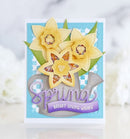 A handmade spring greeting card featuring three yellow daffodil flowers layered over a gray watering can, with the word 'Spring' in purple script and a purple banner reading 'Bright Spring Wishes'. The card is set against a light blue and white checkered background with green leaves, photographed on a white surface with a soft, blurred white floral background.