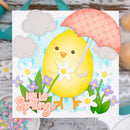A square handmade paper card featuring a cute yellow chick holding a pink plaid umbrella, surrounded by white daisies, purple flowers, and green leaves. The card has 'Hello Spring' written in pink script at the bottom left and is displayed on a rustic wooden surface with craft supplies.